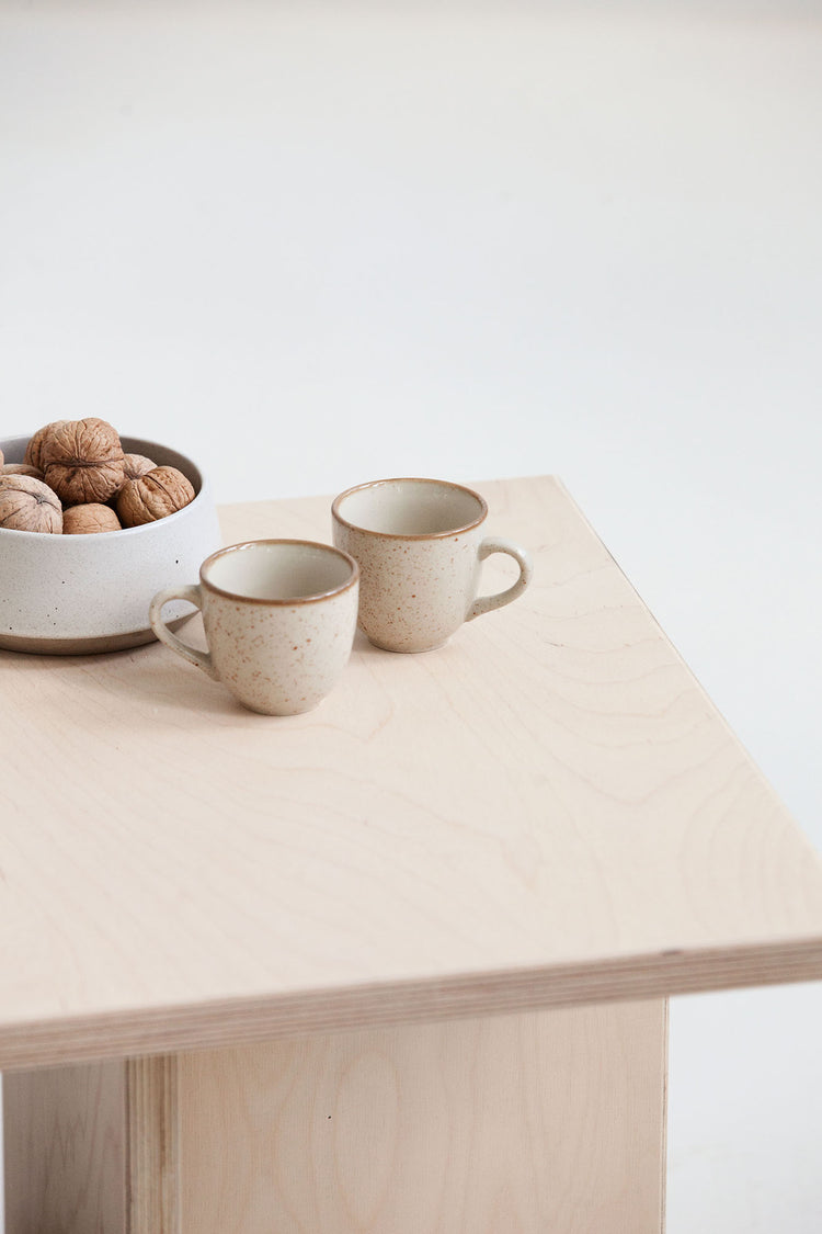 Two ceramic mugs on a light wooden table with a bowl of walnuts.