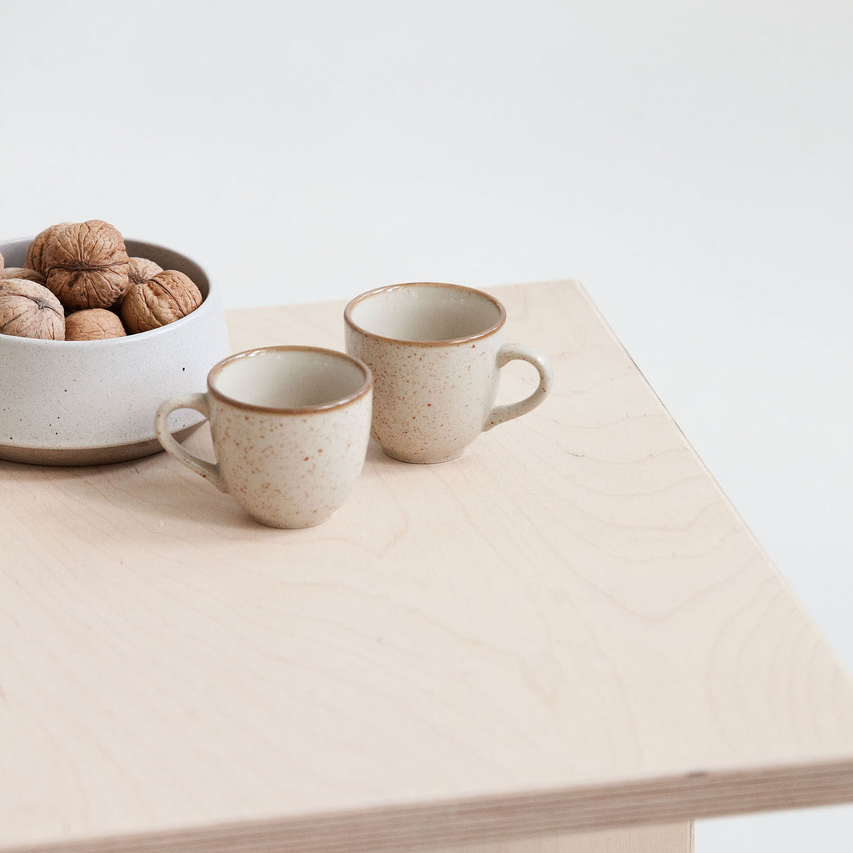 Two ceramic mugs on a light wooden table with a bowl of walnuts.