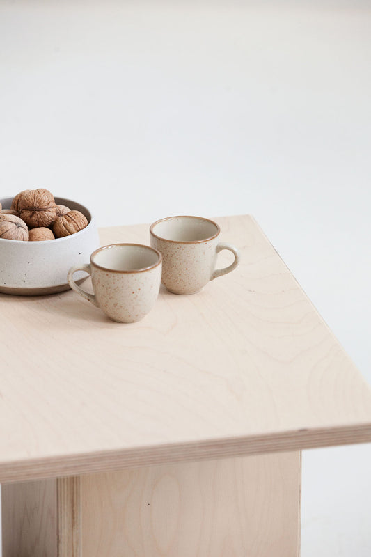 Two ceramic mugs on a light wooden table with a bowl of walnuts.