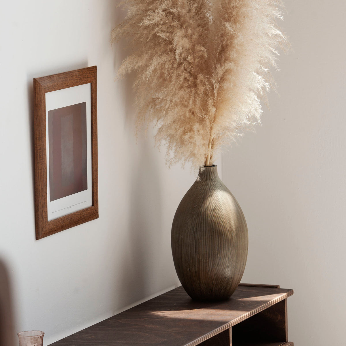 Decorative vase with dried pampas grass on a wooden shelf against a white wall.