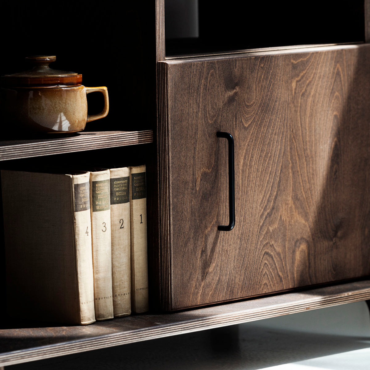 Wooden cabinet with books and a teapot on a shelf against a gray wall.